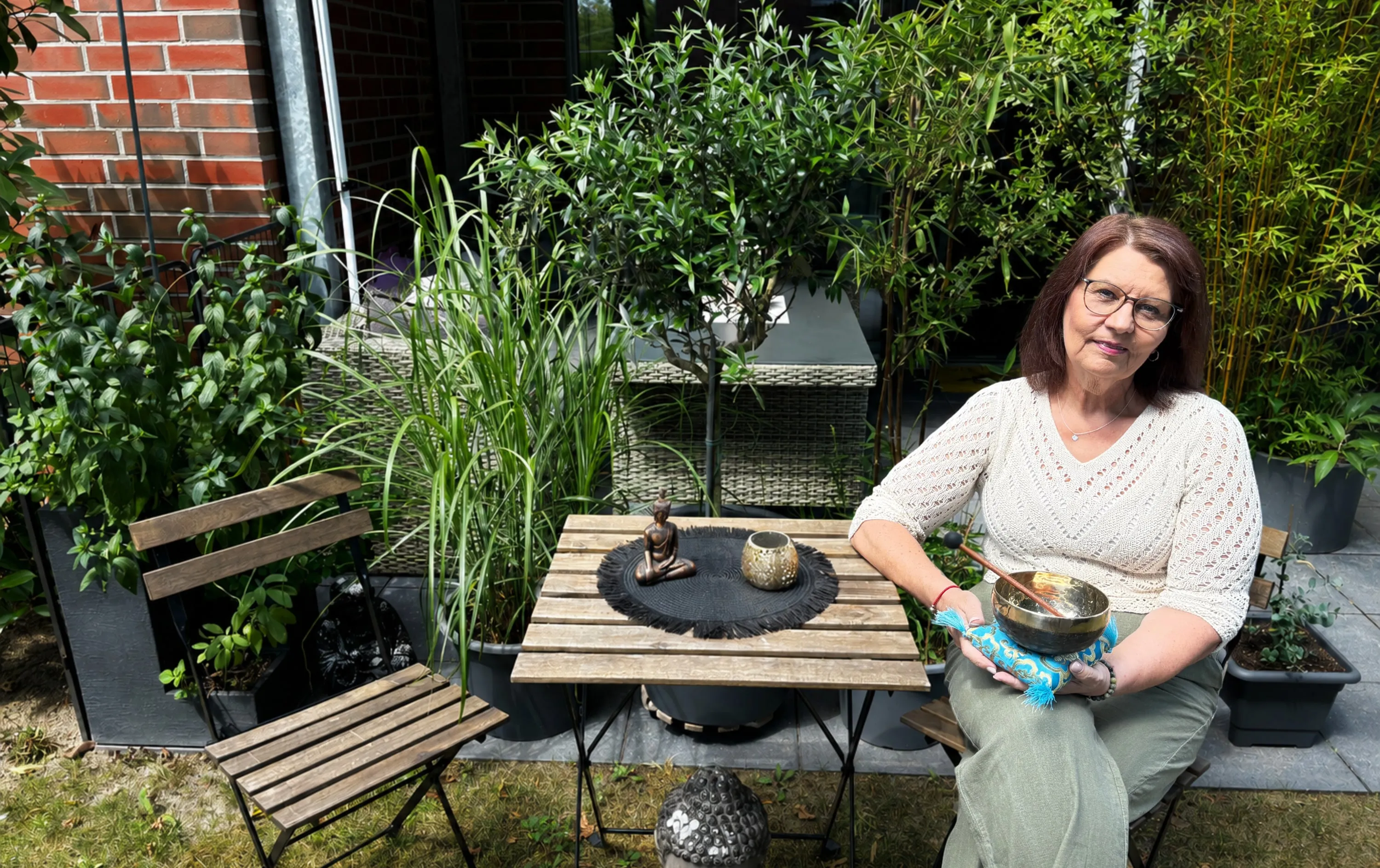 Annette Pitcher sitzt mit einer Klangschale in den Händen auf ihrer Terrasse, umgeben von Grün
